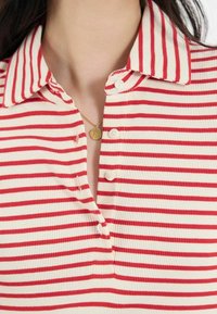 Polo à rayures rouges et blanches, côtelé, avec un col, featuring une patte de boutonnage et un collier avec un pendentif en or.