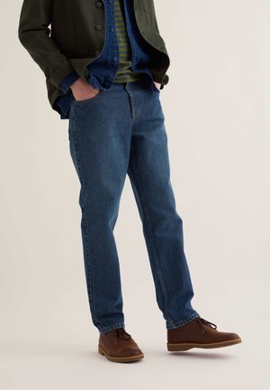 Ein Mann trägt blaue Jeans, braune Lederschuhe, ein gestreiftes Hemd, eine Jeansjacke und einen dunkelgrünen Blazer und steht mit den Händen in den Taschen.