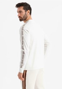 Muž s vousy v bílém dlouhém rukávu tričku se hnědým textem na rukávech a bílých kalhotách s hnědými bočními pruhy, který se dívá přes rameno.