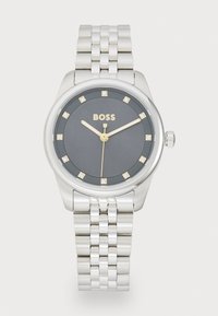Montre-bracelet en argent avec un cadran rond gris, des accents dorés et huit marqueurs ressemblant à des diamants. Elle possède un boîtier en acier inoxydable et une couronne.