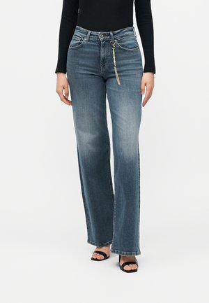 Kvinde iført falmede blå vidde-ben jeans med en dekorativ nøglekæde, sort langærmet top og sorte åbne tå-hæle.