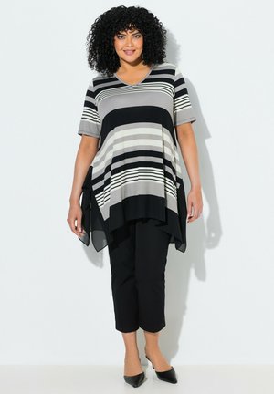 Frau mit lockigem Haar, die eine schwarz-grau-weiß gestreifte asymmetrische Tunika, schwarze verkürzte Hosen und schwarze spitze High Heels trägt.