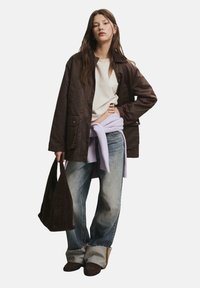 Braune, gesteppten Jacke über einem cremefarbenen Oberteil, kombiniert mit hellblauen, weitem Jeans. Ein lavendelfarbenes Sweatshirt ist um die Taille gebunden. Hält eine dunkle Tasche.