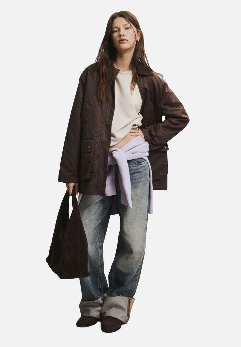 Braune, gesteppten Jacke über einem cremefarbenen Oberteil, kombiniert mit hellblauen, weitem Jeans. Ein lavendelfarbenes Sweatshirt ist um die Taille gebunden. Hält eine dunkle Tasche.