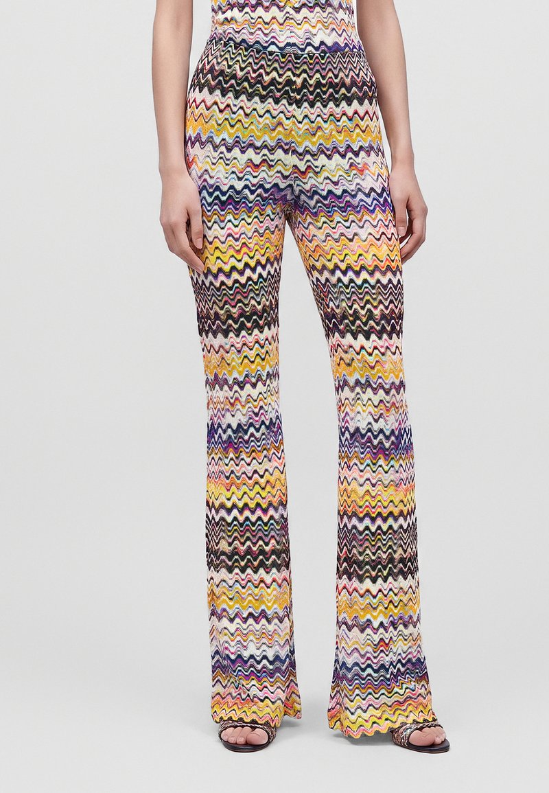Femme portant un pantalon large à motif zigzag multicolore et des sandales noires à bout ouvert sur un fond uni.