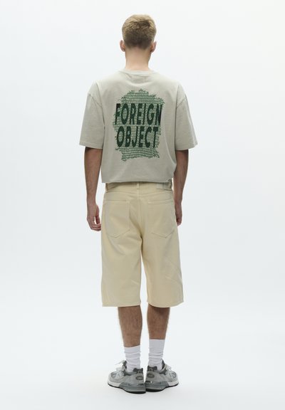 Młody mężczyzna stoi tyłem, ubrany w jasnobeżowe spodenki, pasującą oversize'ową koszulkę z zielonym napisem "FOREIGN OBJECT", białe skarpetki i szare tenisówki.