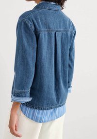 Veste en denim bleu foncé avec un col boutonné, des manches trois-quarts et un dos plissé. Portée par-dessus une chemise à rayures bleu clair.