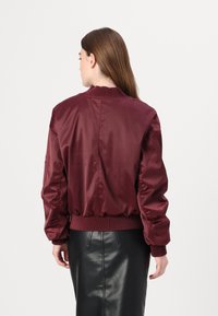 Giacca bomber in raso bordeaux con colletto e polsini a costine, caratterizzata da un design cropped. Indossata sopra una gonna di pelle nera.