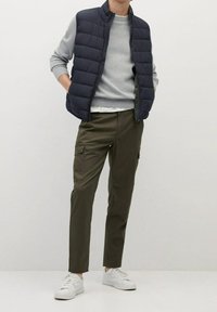 Marineblaue gepolsterte Weste über einem grauen Pullover, kombiniert mit olivgrünen Cargo-Hosen und weißen Sneakern. Die Weste hat einen hohen Kragen und einen Reißverschluss.