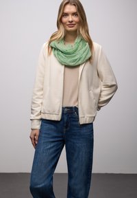 Veste bomber couleur crème avec texture côtelée, associée à un haut beige et un jean bleu. Écharpe verte à motifs drapée autour du cou.
