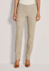 Vrouw in beige, met een patroon, slim-fit broek en bruin, puntige hakken, staand voor een effen lichte achtergrond.