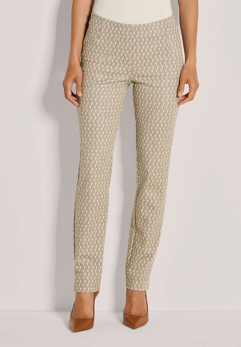 Vrouw in beige, met een patroon, slim-fit broek en bruin, puntige hakken, staand voor een effen lichte achtergrond.