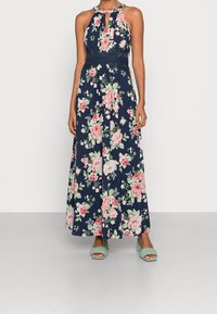 Maxime luettelo tummansinisessä värissä, jossa on vaaleanpunaisia kukkia, niskalenkillä, venyvässä smokkibodissa ja hulmuavassa hameessa. Yhdistetty vihreisiin liukuviin sandaaleihin.