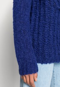 Pletený svetr v tmavě modré barvě, s texturovaným vzorem a vroubkovanými manžetami. Materiál působí měkce a načechraně. Ležérní design.