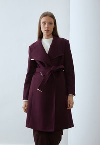 Cappotto in misto lana borgogna con ampi risvolti, vita con cintura e dettagli in metallo argentato. Maniche lunghe e lunghezza sotto il ginocchio. Texture liscia.
