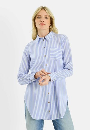 Mujer con camisa de botones a rayas verticales azules y blancas con bolsillo en el pecho, ajustándose su pulsera de oro, vistiendo vaqueros azul claro.