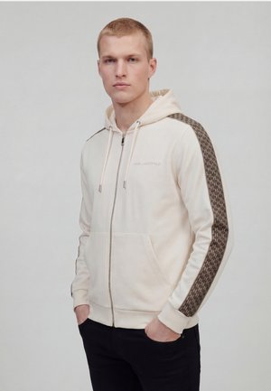 Young man wearing a cream zip-up hoodie with brown patterned stripes on sleeves and black pants, standing with hands in pockets.