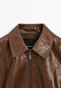 Brown leather jacket with a collared neckline, featuring a central zipper and smooth texture. Label visible inside collar area.