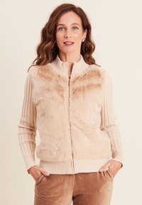 Gilet zippé beige avec devant en fausse fourrure, manches tricotées à côtes et ourlet. Comprend des poches latérales et un col haut. Texture douce partout.