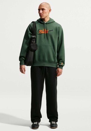 Mann trägt grünen Nike-Hoodie, schwarze Hose und schwarze Sneakers, trägt eine schwarze Umhängetasche, steht vor schlichtem weißem Hintergrund.