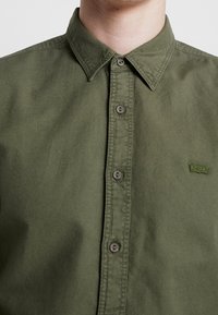 Olijfgroene katoenen overhemd met knopen, voorzien van een spreidkraag, bijpassende knopen en een klein groen Levi's-logo.