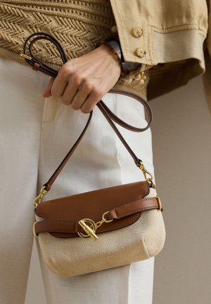 Mână ținând o geantă crossbody bej și maro cu închizătoare aurie, purtând un pulover bej texturat și pantaloni deschiși la culoare.