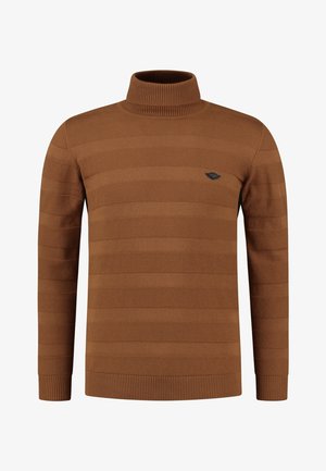 Man draagt een roestkleurige coltrui, donkere geruite broek en tan suède laarzen, staat met ontspannen handen.