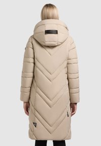 Beige polstret lang frakke med chevron-syning, stor hætte og sort logo-tag. Glat tekstur og let skær. Side lommer synlige.