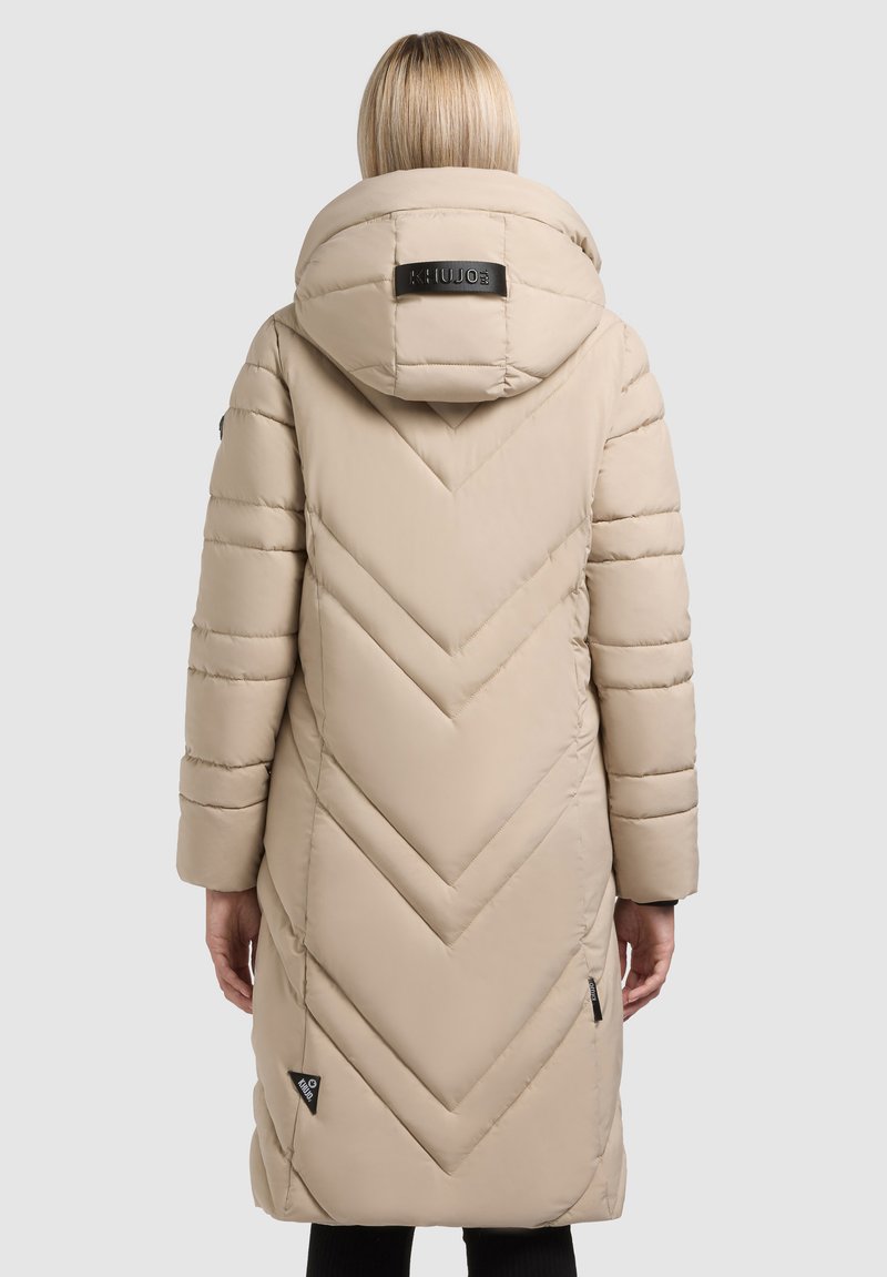 Beige polstret lang frakke med chevron-syning, stor hætte og sort logo-tag. Glat tekstur og let skær. Side lommer synlige.