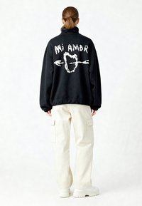 Pull noir en molleton avec col montant et texte blanc imprimé « Mi Amor » ainsi que motif graphique au dos ; associé à un pantalon cargo crème et des baskets blanches.