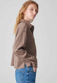 Brown long-sleeve shirt with a collar, featuring a front pocket and side slits, paired with blue jeans. Soft texture, relaxed fit.
