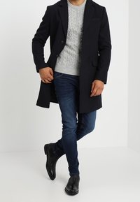 Uomo con cappotto nero, maglione grigio a trecce, jeans blu scuro e scarpe di pelle nere, in piedi con una gamba incrociata davanti.