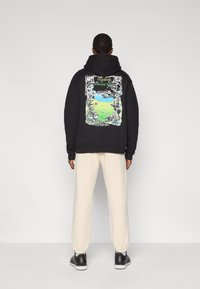 Μαύρο φούτερ με κουκούλα από βαμβάκι με πολύχρωμη γραφική εκτύπωση στην πλάτη που απεικονίζει ένα τοπίο, σε συνδυασμό με κρεμ παντελόνια φόρμας και μαύρα αθλητικά παπούτσια.