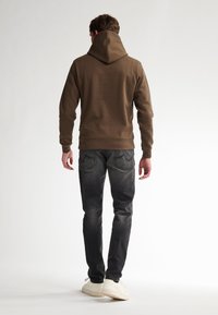 Bruine hoodie met een gladde textuur, gecombineerd met donkergrijze jeans en witte sneakers, gezien van achteren tegen een lichte achtergrond.
