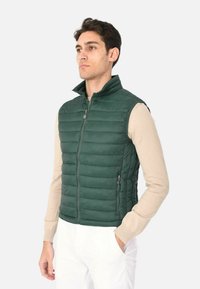 Gilet trapuntato verde con colletto alto, zip frontale e tasche laterali, indossato sopra una maglietta beige a maniche lunghe, presentato su uno sfondo bianco.