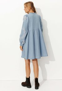 Robe en denim bleu clair avec des manches longues ornées de broderies, une jupe froncée et un col rond. Associée à des bottines chelsea marron foncé.