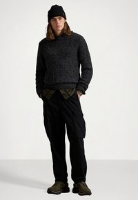 Σκούρο γκρι πλεκτό πουλόβερ με ψηλό γιακά, πράσινο καρό πουκάμισο από κάτω, μαύρο παντελόνι cargo και σκούρα αθλητικά παπούτσια, ολοκληρωμένα με μαύρο σκουφί.