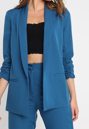 Frau trägt einen blauen Blazer mit gerafften Ärmeln über einem schwarzen, bauchfreien Top und passenden blauen hoch taillierten Hosen vor weißem Hintergrund.