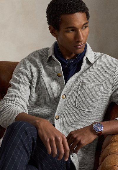 Νέος άνδρας καθισμένος σε καφέ δερμάτινη καρέκλα, φοράει γκρι πλεκτή ζακέτα, σκούρο μπλε φουλάρι με πουά, ριγωτό παντελόνι και μπλε ρολόι χειρός.