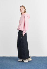 Sudadera con capucha rosa, falda negra de corte holgado con bolsillos y zapatillas beige. Diseño simple, material de algodón y marca sutil en la falda.