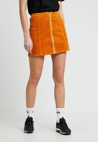 Minifalda de pana naranja con cierre frontal, dos bolsillos laterales y un diseño ajustado. Combinada con zapatos negros y calcetines blancos con logo.