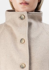 Gros plan d'un manteau en laine beige avec un col haut et trois grands boutons ronds fermés, porté par une personne à la peau claire et aux cheveux blonds.