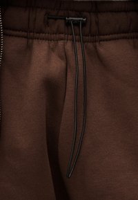 Καφέ ύφασμα με σούρα στη μέση και μαύρο κορδόνι δεμένο σε θηλιά στο κέντρο μπροστά.