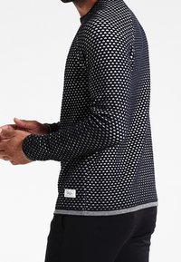 Chemise à manches longues noire avec un motif triangulaire géométrique blanc. Matière douce avec des poignets côtelés et une étiquette de marque à l'ourlet.