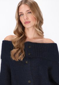Sötétkék, válltól elálló kardigán textúrált kötésben, öt sötét gombbal az elülső részen, hosszú ujjakkal és bordázott nyakkivágással.