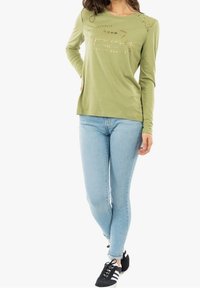 Oliivinvihreä pitkähihainen paita, jossa on pitsikoristeet olkapäillä, kultateksti "Superdry NYC", yhdistettynä vaaleansinisiin kapealahkeisiin farkkuihin ja mustiin lenkkareihin.