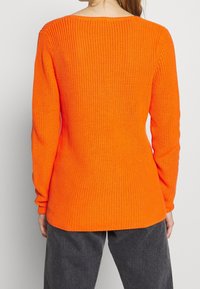 Oranger Strickpullover mit Ripptextur, langen Ärmeln und lockerer Passform. Die Rückansicht zeigt einen runden Halsausschnitt und ein auffälliges Muster aus Maschen.
