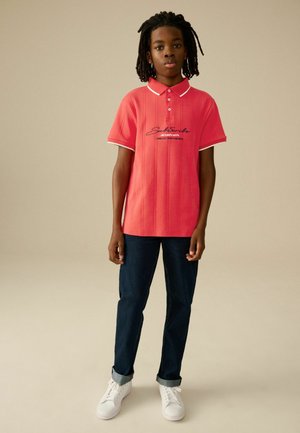 Polo de manga corta en rosa brillante con cuello y acentos de manga blancos, con impresión de texto en negro. Combinado con jeans oscuros y zapatillas blancas.