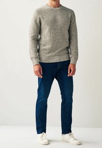 Homme portant un pull gris texturé, un jean bleu foncé et des baskets blanches, debout devant un fond clair uni.