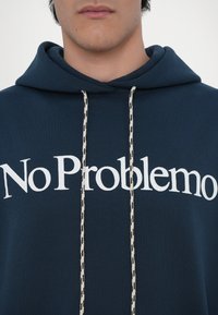 Námořnická modrá mikina vyrobená z bavlny, zdobená velkým bílým nápisem "No Problemo" a krémovými tkaničkami s černými vzorovými akcenty.